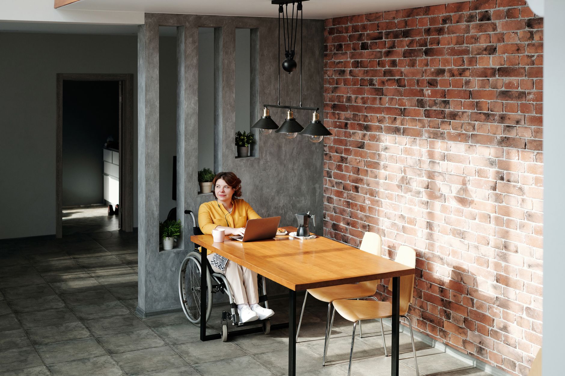 A woman in a wheelchair works on a laptop at a long wooden table beside a brick wall, with a coffee pot and mug nearby